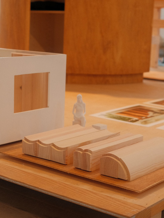 Træmodeller og miniaturefigur på bord ved siden af arkitekturmodel.