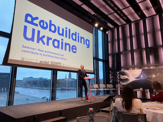 Oplægsholder står på en scene foran et stort lærred med teksten “Rebuilding Ukraine” under et seminar.