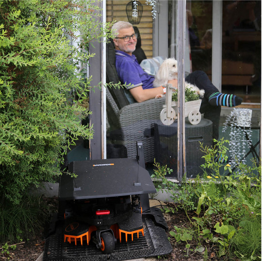 Robotplæneklipper i haven foran en person, der sidder indendørs med en hund.