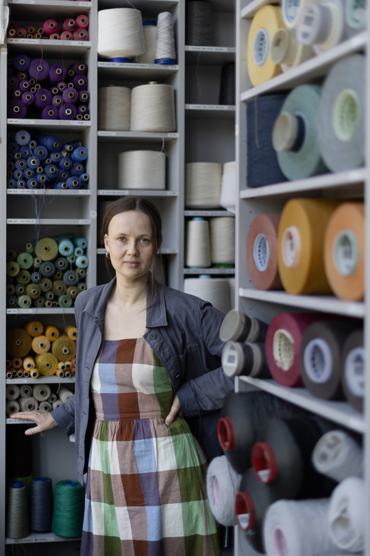 The image is portraying Trine in front of shelves filled with cones of yarn. She is wearing a blue jacket and a checkered dress in red, green, blue and white.