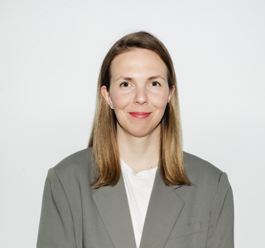 Professional portrait of a woman with shoulder-length light brown hair wearing a grey blazer and white top against a light background.
