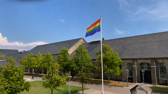 Et regnbueflag på flagstang vajer over solbeskinnet campus
