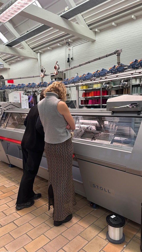 Two people examine an industrial knitting machine surrounded by colorful yarn.