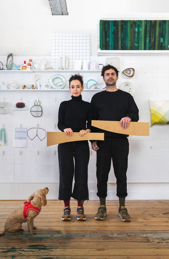 Two people in black hold wooden pieces in a design studio as a dog looks on.