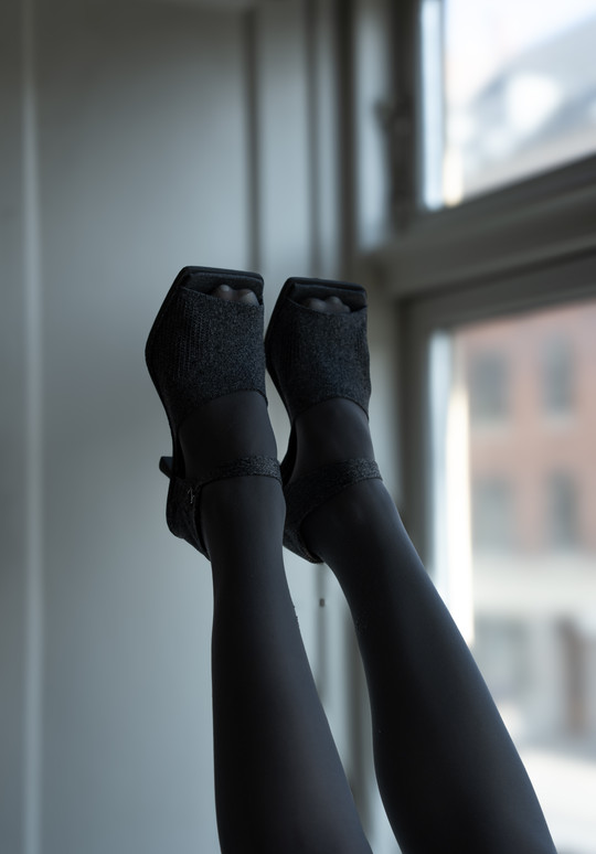 Feet in black open-toe high-heeled dance shoes raised in front of a window.
