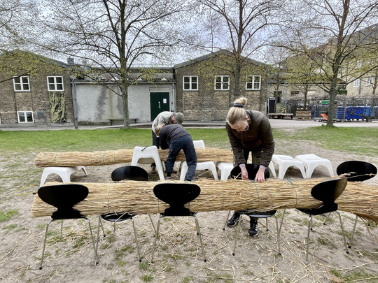 Tre personer arbejder med at binde lange stråbundter sammen udendørs foran en murstensbygning.
