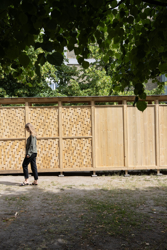 Kvinde går forbi en trækonstruktion med glatte og flettede paneler under grønne træer.