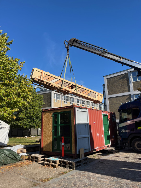 En kran løfter en lang trægitterkonstruktion over en rød container ved en industribygning.
