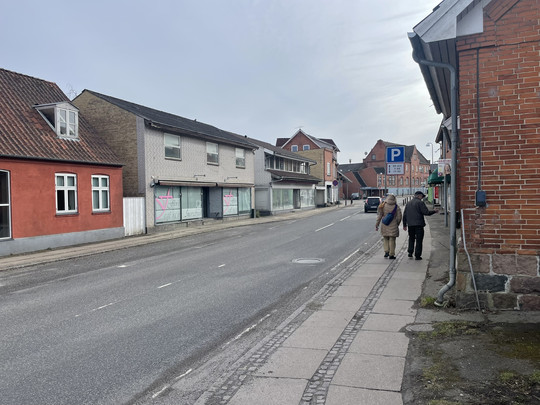 To personer går langs en stille bygade med ældre murstenshuse og tomme butikslokaler.