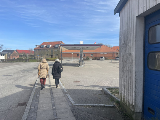 To personer går på fortovet forbi en åben plads med en ladestander og røde murstensbygninger.