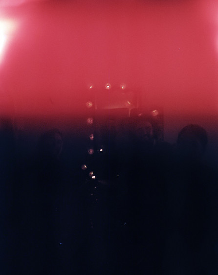 Photograph of a group of people sitting around a table. The image is obstructed by a black and red patch. Only what appears to be a string of lights and the shilouettes of three heads in the center are visible.  