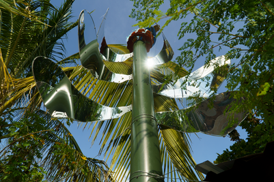 Skulpturel, palmelignende metalinstallation på høj mast, der reflekterer sollys mellem grønne træer og blå himmel.