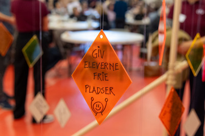 Orange skilt med teksten "Giv eleverne frie pladser" hænger foran en sløret workshop.