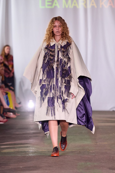 Model går på catwalk i en lys kappe med lilla frynse- og patchworkdetaljer, lilla for og rødsorte sko