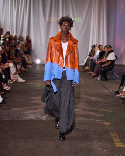 Model går på catwalk i en orange læderjakke med lyseblå afslutning, vide mørke bukser og hvid top.