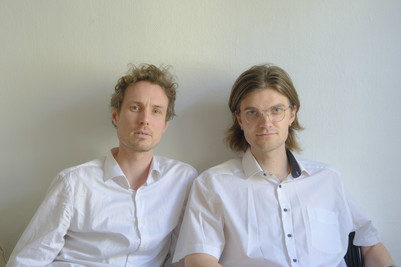 Two men in white shirts sitting side by side against a light wall.