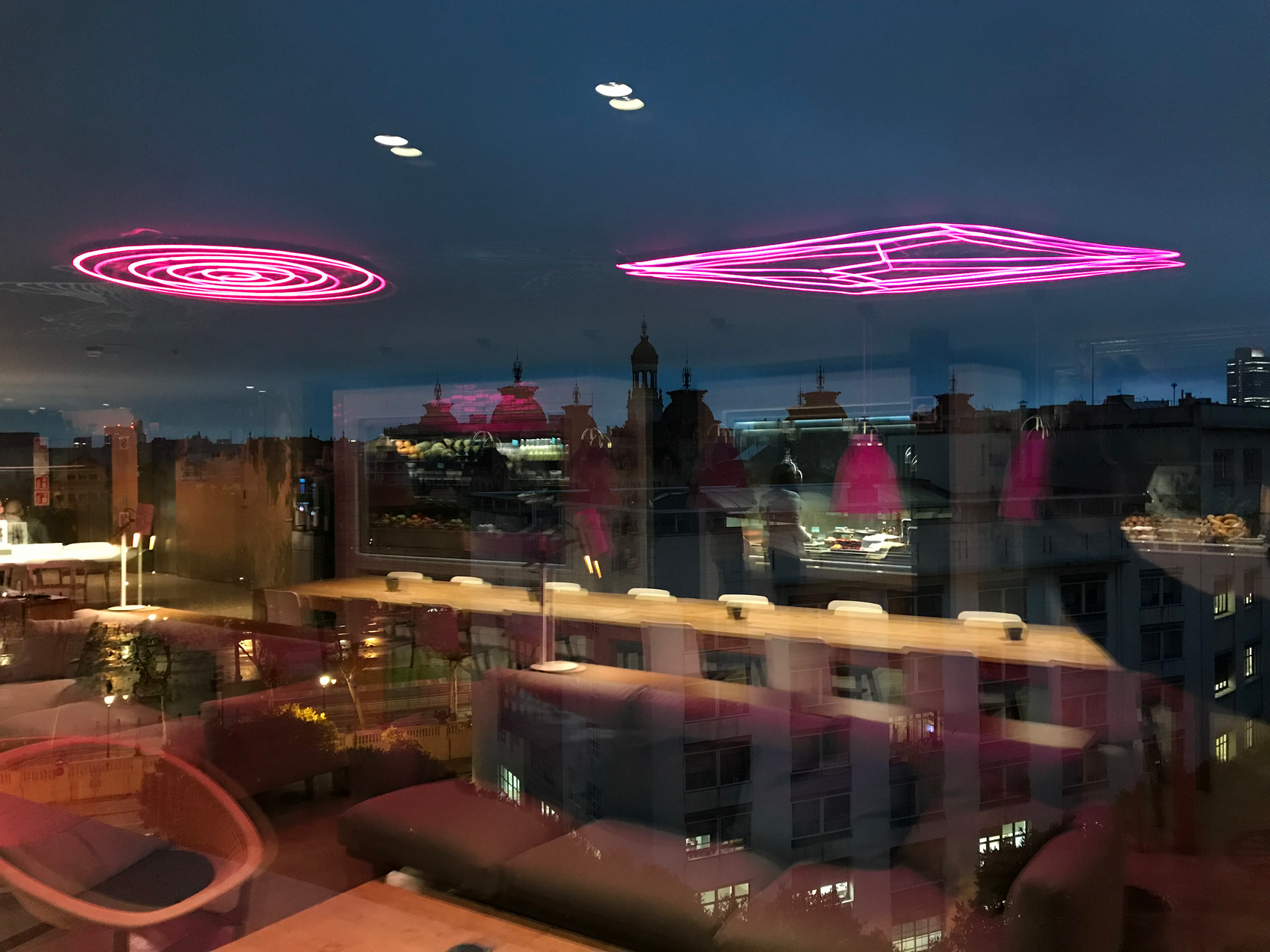  Neonlys i loftet spejler sig i glas med udsigt over byens tage i skumringen.