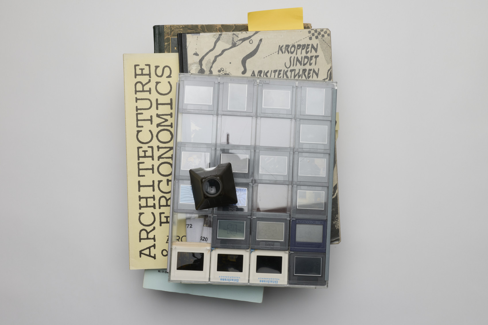 A stack of archival material rests on a light surface, topped with a clear sheet of photo slides arranged in a grid. A small black slide viewer rests on top, and sticky notes stick out from the pages. 