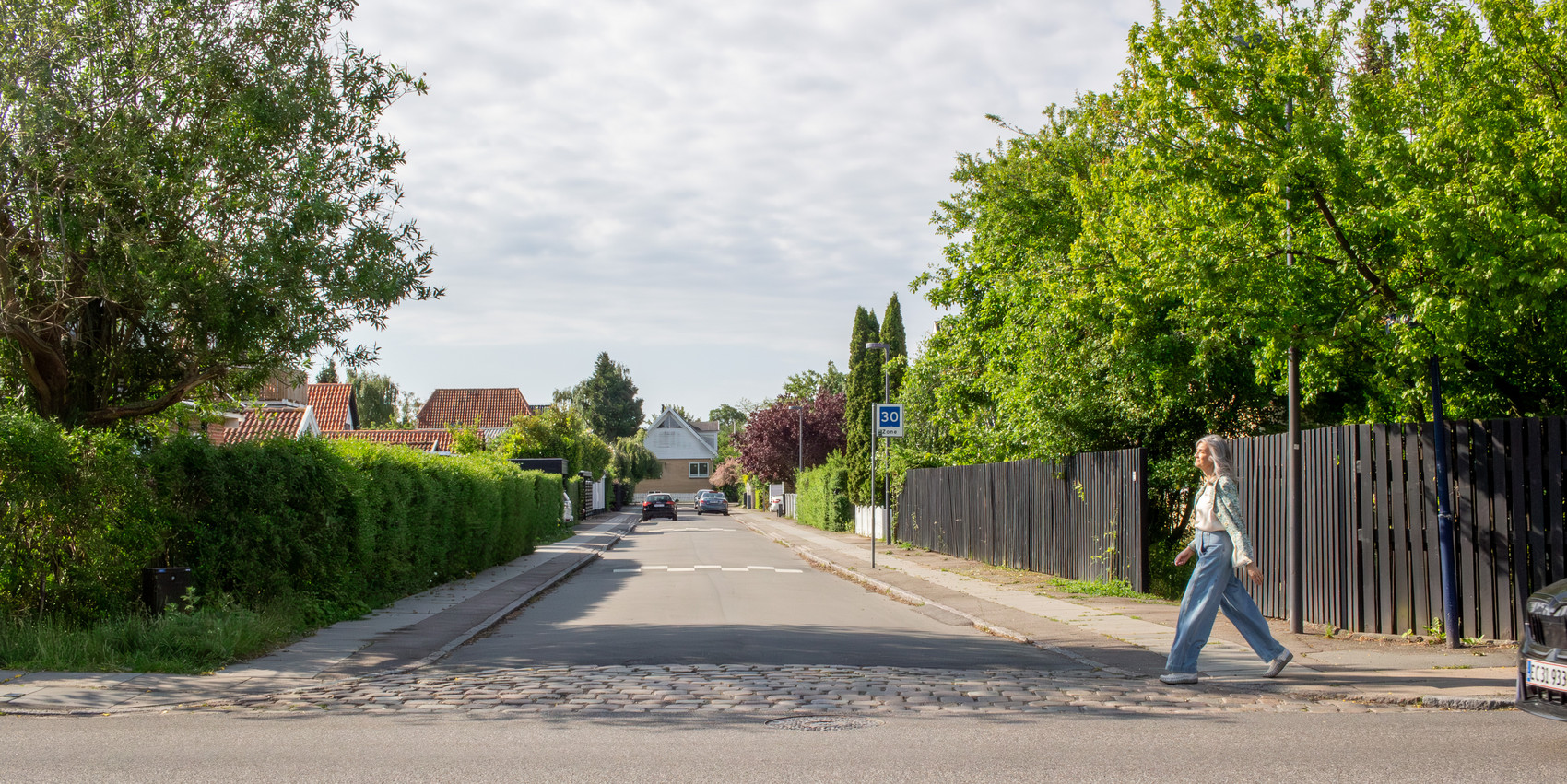 A woman crossing a street, green trees on each sides and houses behind hedges