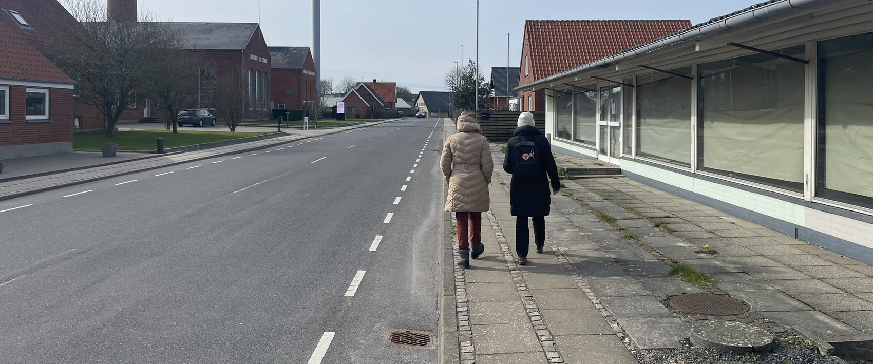 To personer går langs fortovet i en mindre by med lave bygninger