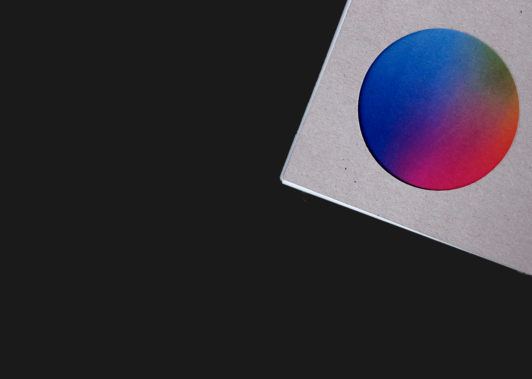 A thick cardboard book cover lies diagonally in the top-right corner against a dark background. A circular cutout in the cover reveals the colorful first inner page beneath, showing a soft gradient of blue, purple, red, and green.