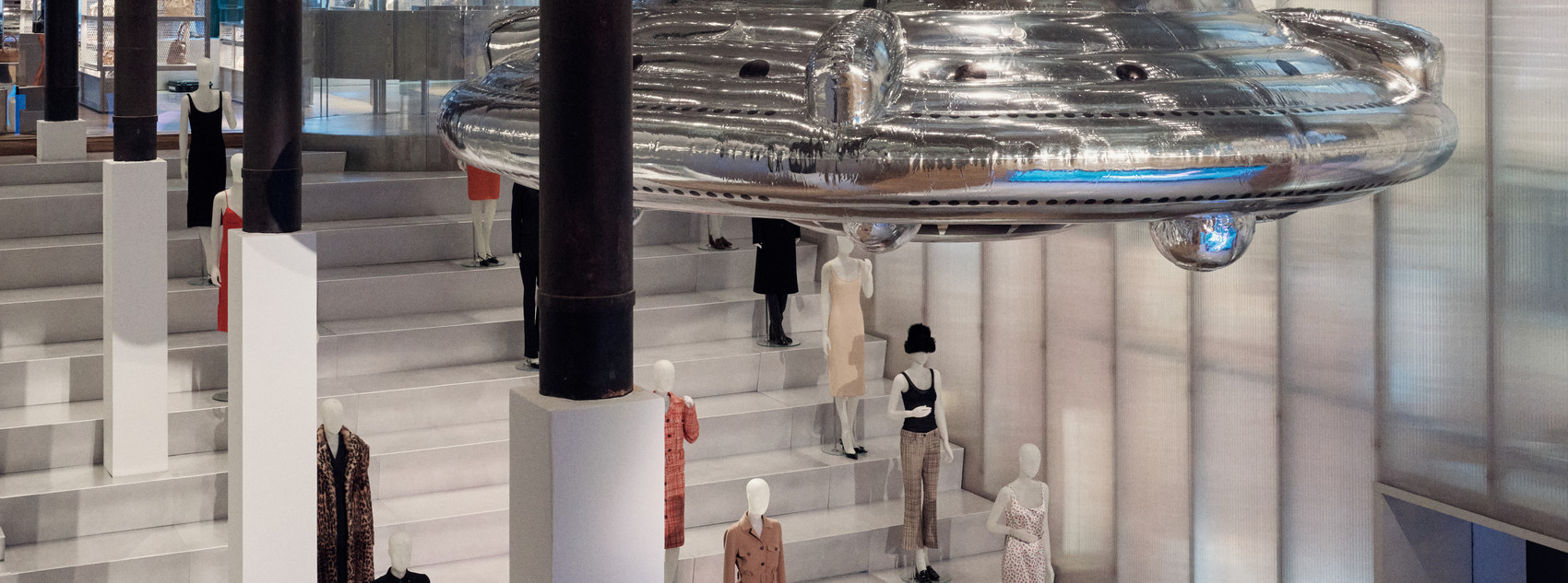 Large silver UFO-like installation hangs above a fashion exhibition.