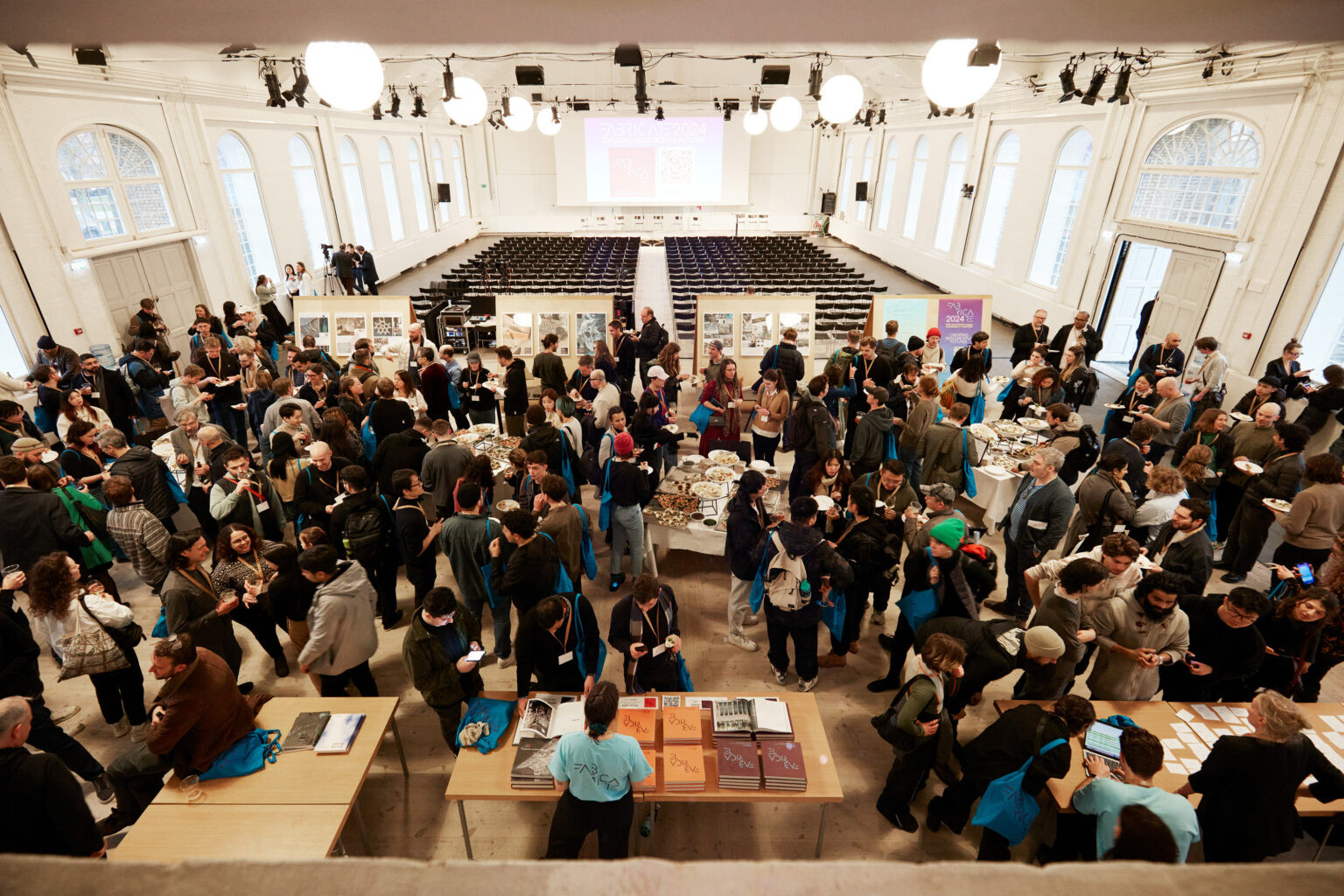 A large crowd networks around buffet tables in a bright conference hall with rows of chairs in the background.