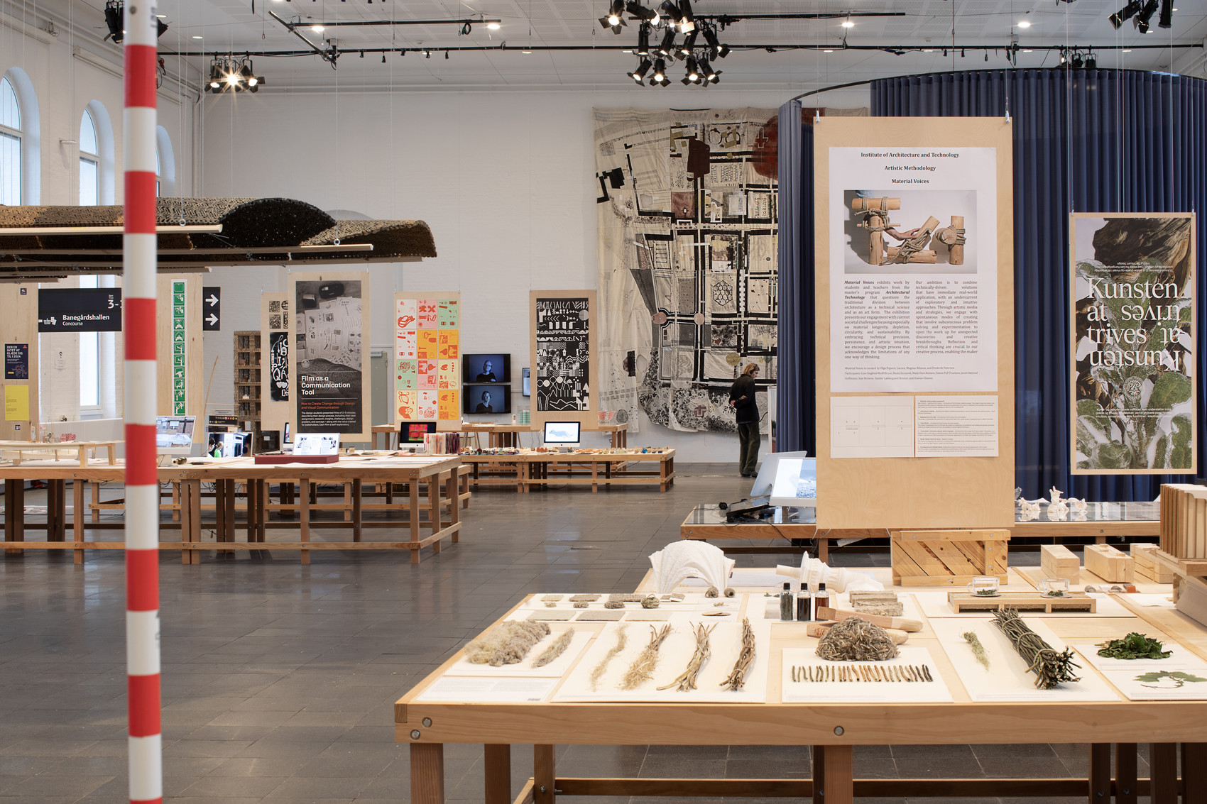 Udstillingssal med træborde, naturmaterialer, modeller og plancher om kunst og design.